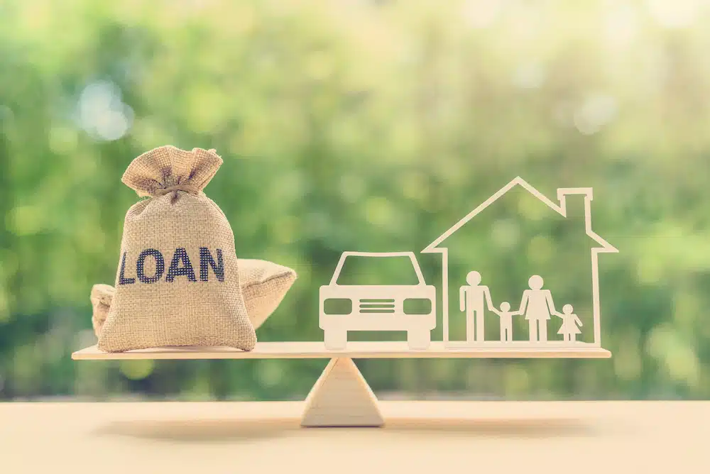 Loan money bag balancing with house, car, and family on wooden seesaw